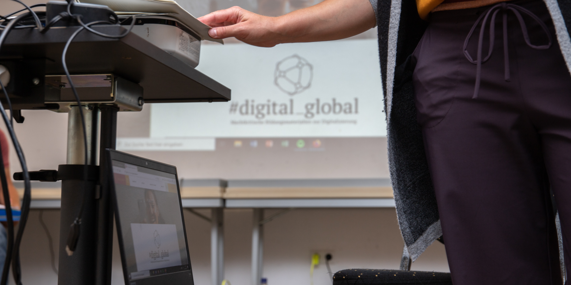 Eine Person steht neben einem Beamer. Neben ihr steht ein Laptop. Aufgerufen ist die Website des Projektes digital global. Im Hintergrund ist das Logo von digital global an der Wand zu sehen.
