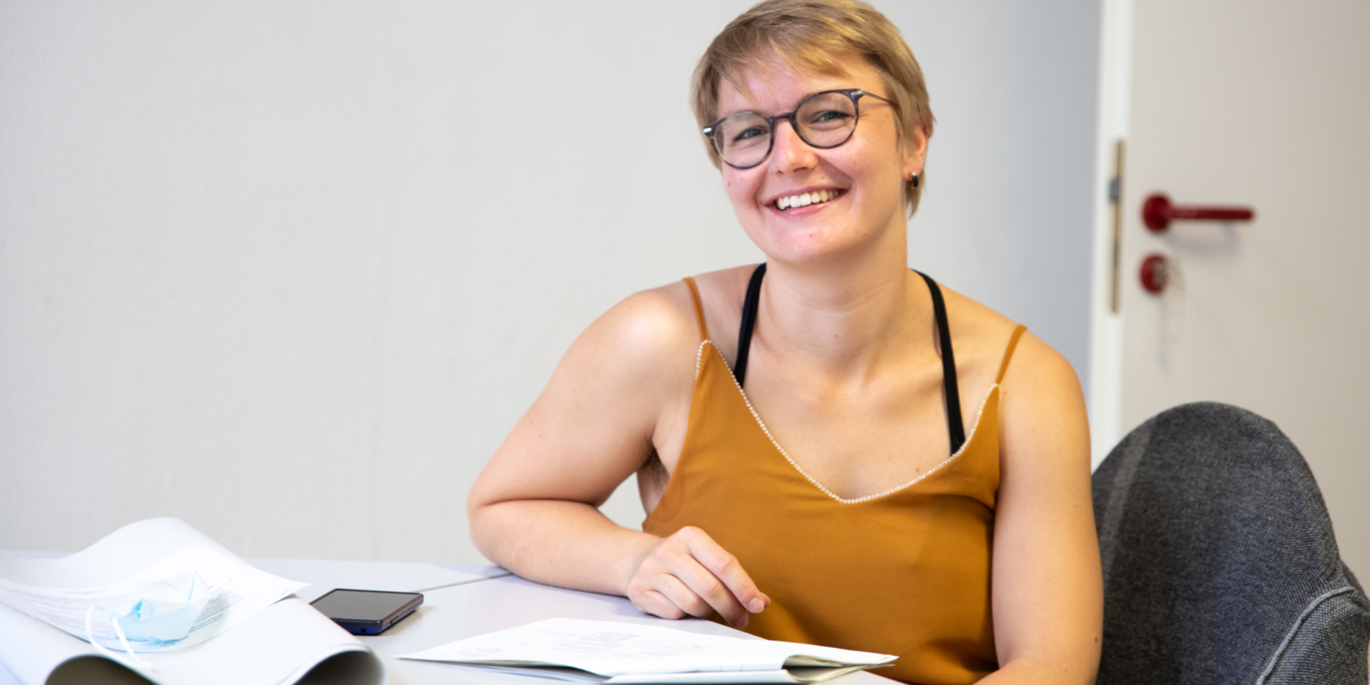 Kollektivmitglied Evelyn sitzt lachend an einem Tisch. Sie trägt eine dunkelbraune Brille und ein orangenes Top.