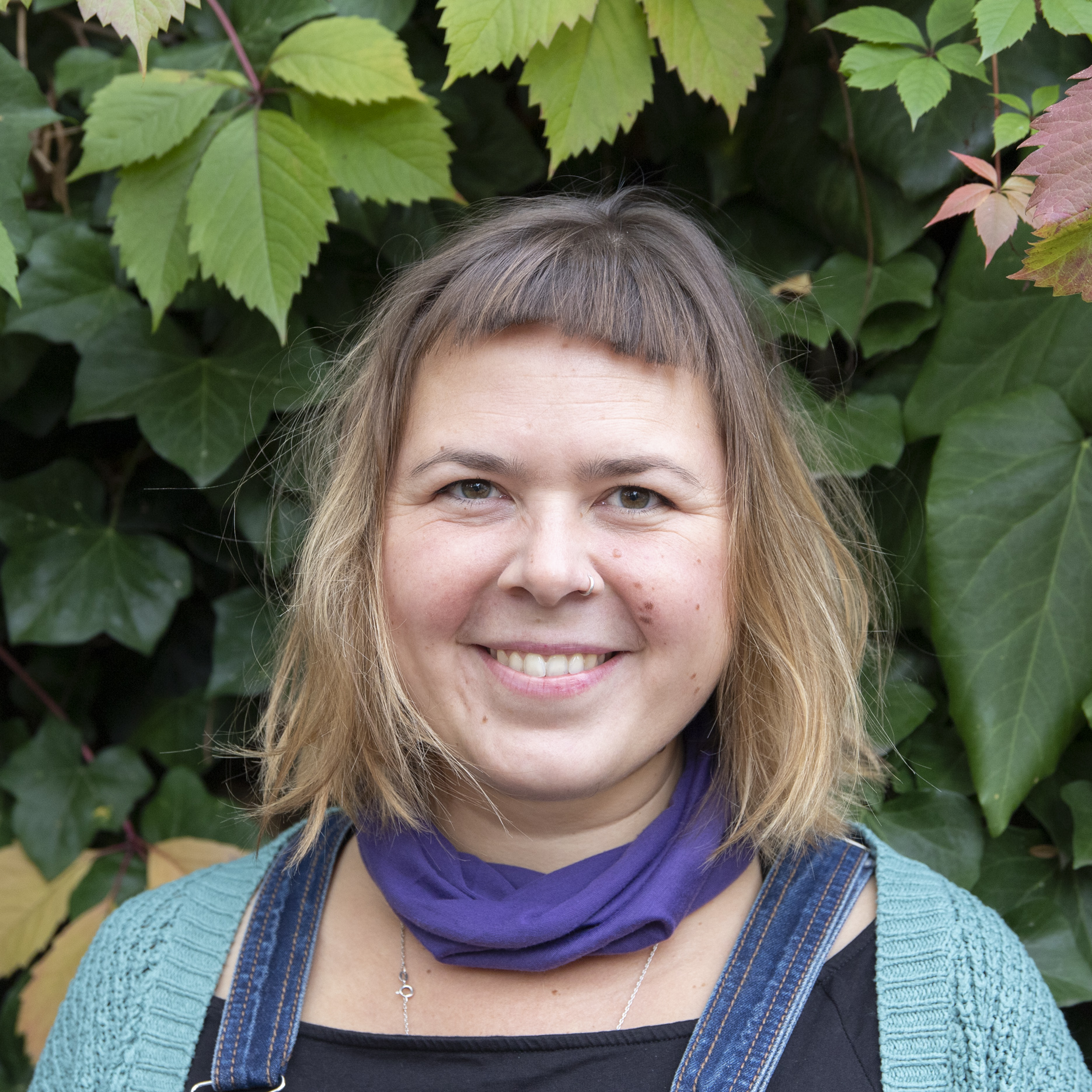 Portrait von Kollektivmitglied Katrin vor einem Busch mit grünen Blättern. Sie hat einen Pony, trägt einen lilanen Schal und eine türkisene Strickjacke und lächelt.