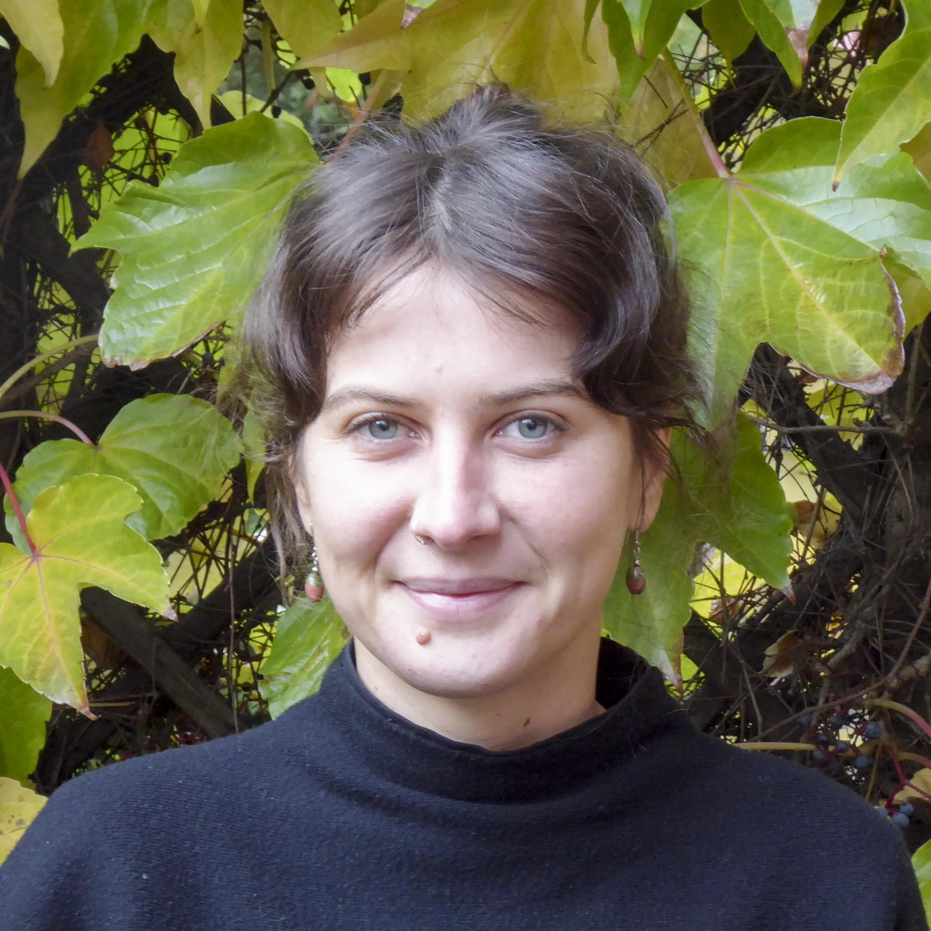 Portrait von Kollektivmitglied Maike vor einem Baum mit grünen und gelben Blättern. Sie trägt einen schwarzen Pullover und orangene Ohrringe und lächelt in die Kamera.