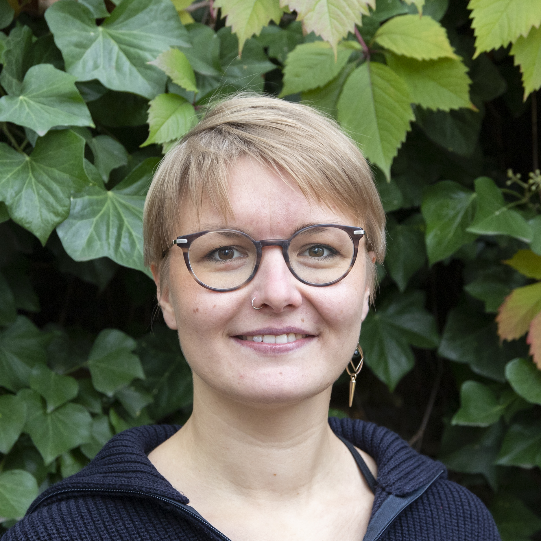 Portrait von Kollektivmitglied Evelyn vor einem Busch mit grünen Blättern. Sie trägt eine dunkelbraune Brille und einen goldenen Ohrring und lächelt.
