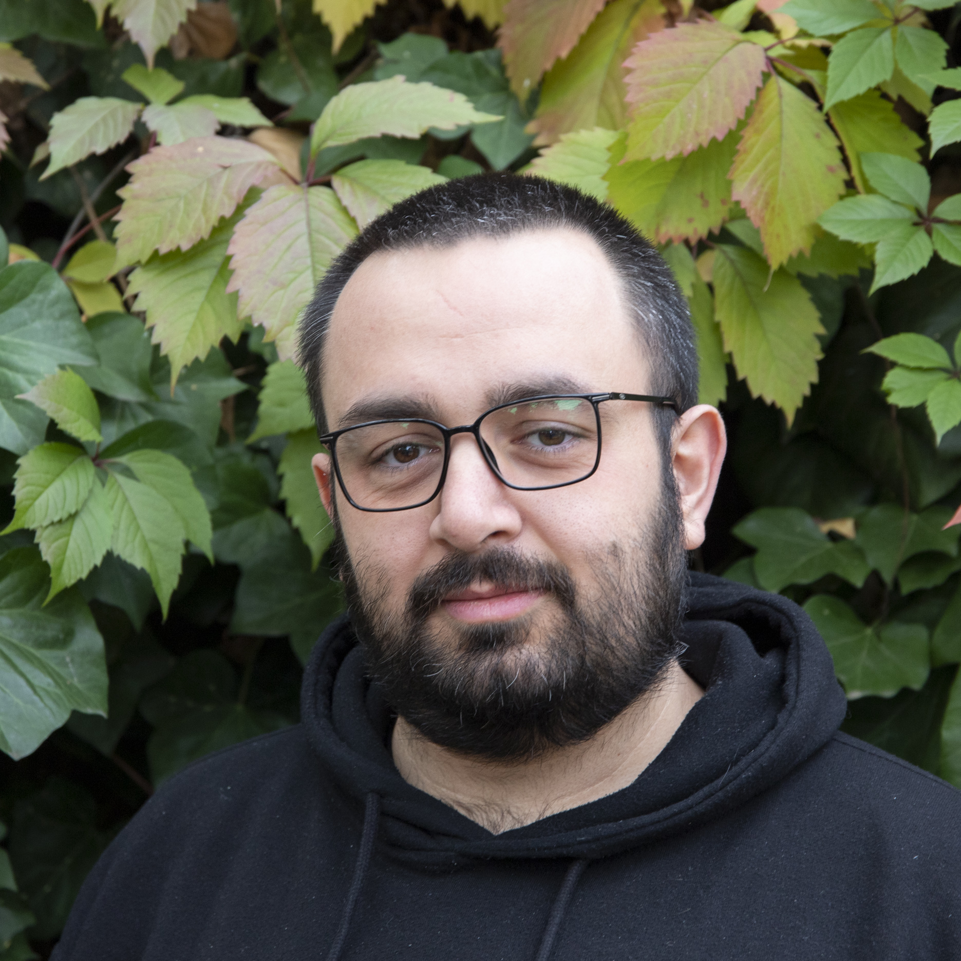 Portrait von Kollektivmitglied Mario vor einem Busch mit grün-roten Blättern. Mario trägt eine schwarze Brille und einen schwarzen Pullover und hat ein Lächeln auf den Lippen.