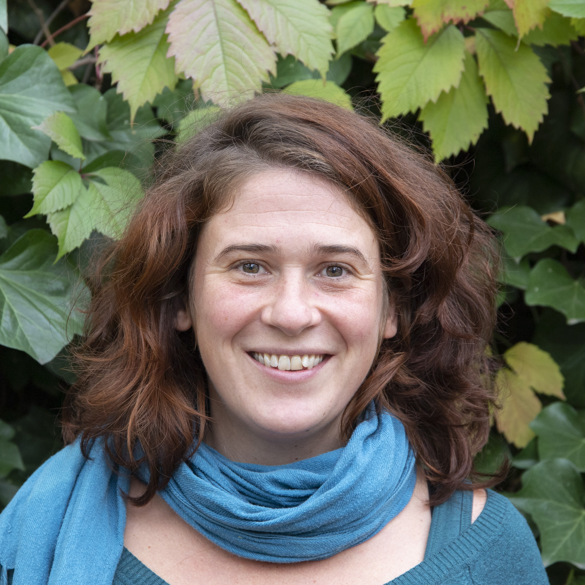 Portrait von Kollektivmitglied Marie vor Efeu mit grünen Blättern. Sie trägt einen türkisenen Schal und türkisenen Pullover und lacht.