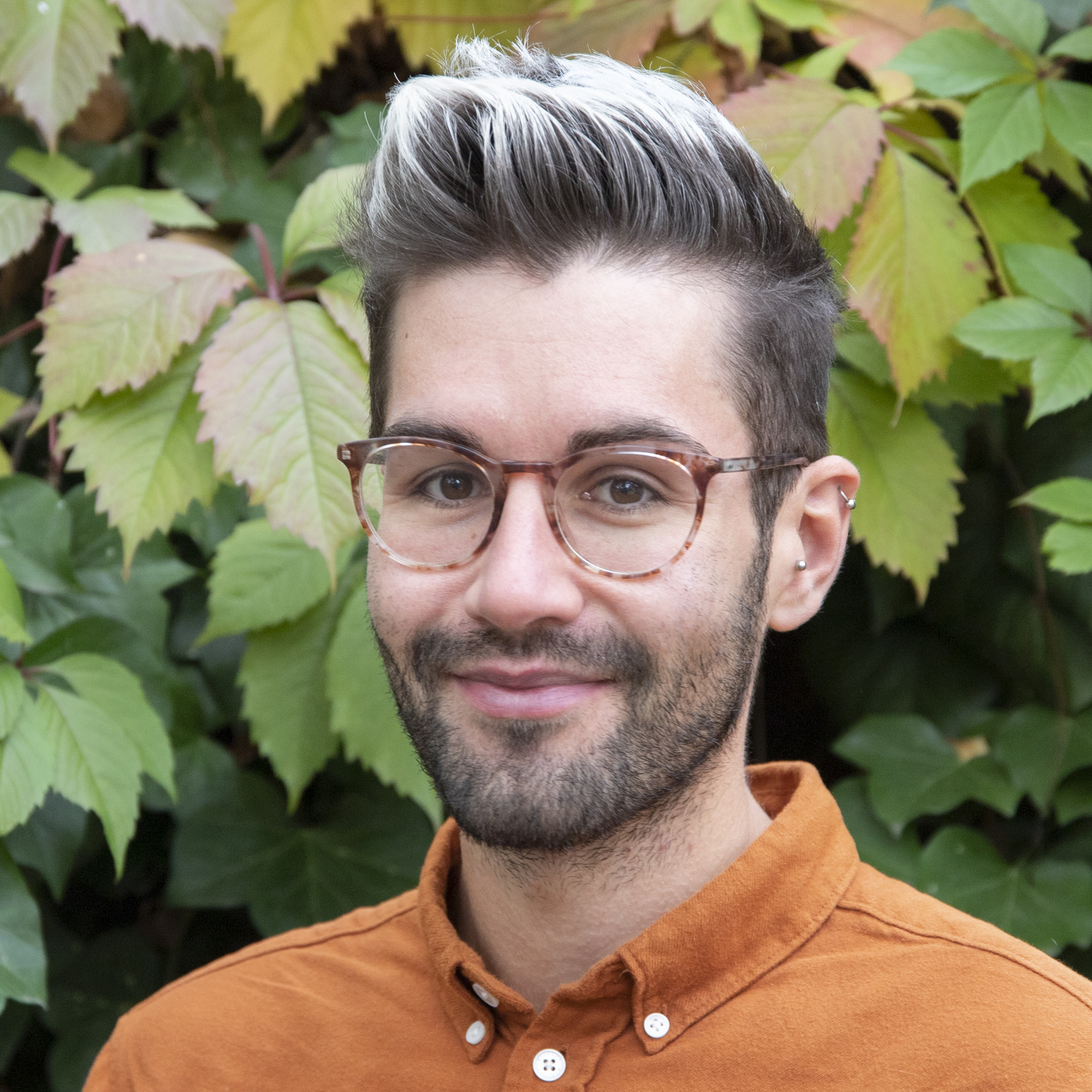 Portrait von Kollektivmitglied Simon vor einem Busch mit grün-roten Blättern. Er trägt eine hellbraune Brille und ein orangenes Hemd und lächelt..