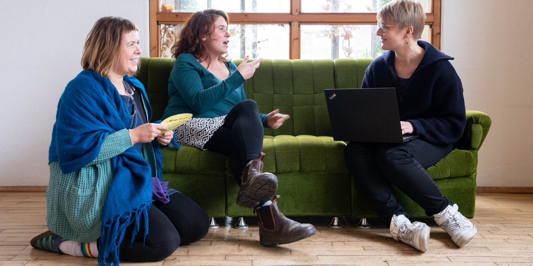 Katrin, Marie und Evelyn vom F3_kollektiv sitzen nebeneinander und diskutieren. Katrin kniet lächelnd auf dem Boden und hält beschriftete Moderationskarten in der Hand. Marie sitzt daneben auf dem Sofa und gestikuliert mit den Händen. Rechts von ihr sitzt Evelyn, ihr zugeneigt, mit einem Laptop auf dem Schoß und lächelt.