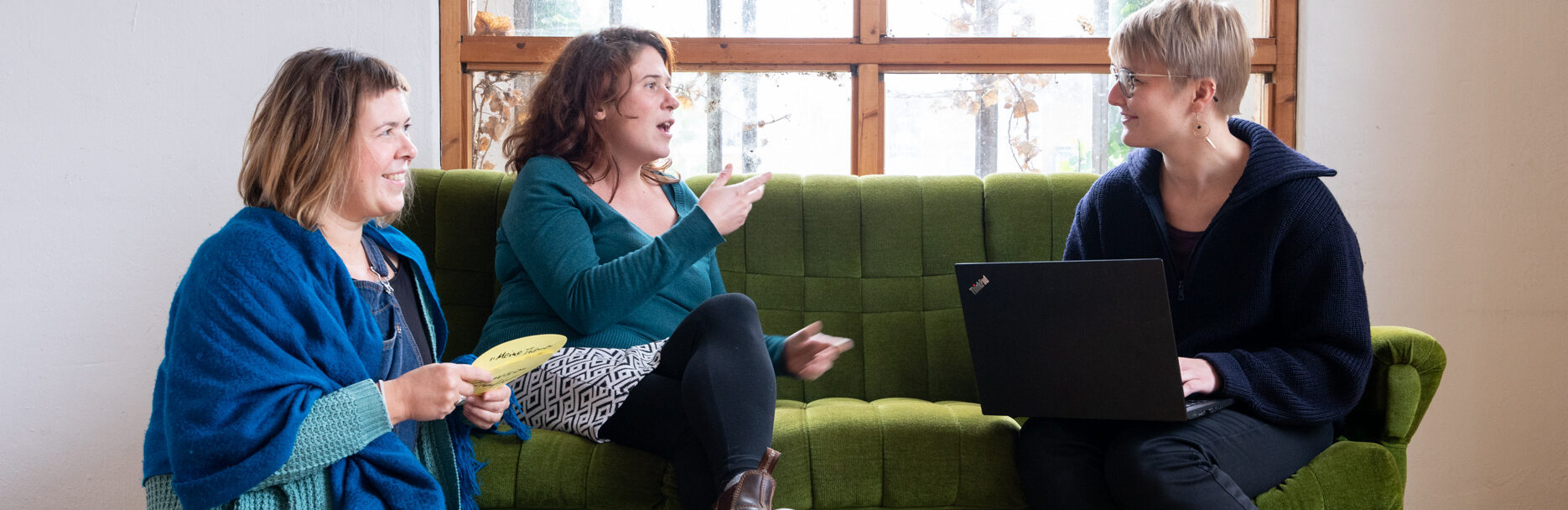 Katrin, Marie und Evelyn vom F3_kollektiv sitzen nebeneinander und diskutieren. Katrin kniet lächelnd auf dem Boden und hält beschriftete Moderationskarten in der Hand. Marie sitzt daneben auf dem Sofa und gestikuliert mit den Händen. Rechts von ihr sitzt Evelyn, ihr zugeneigt, mit einem Laptop auf dem Schoß und lächelt.