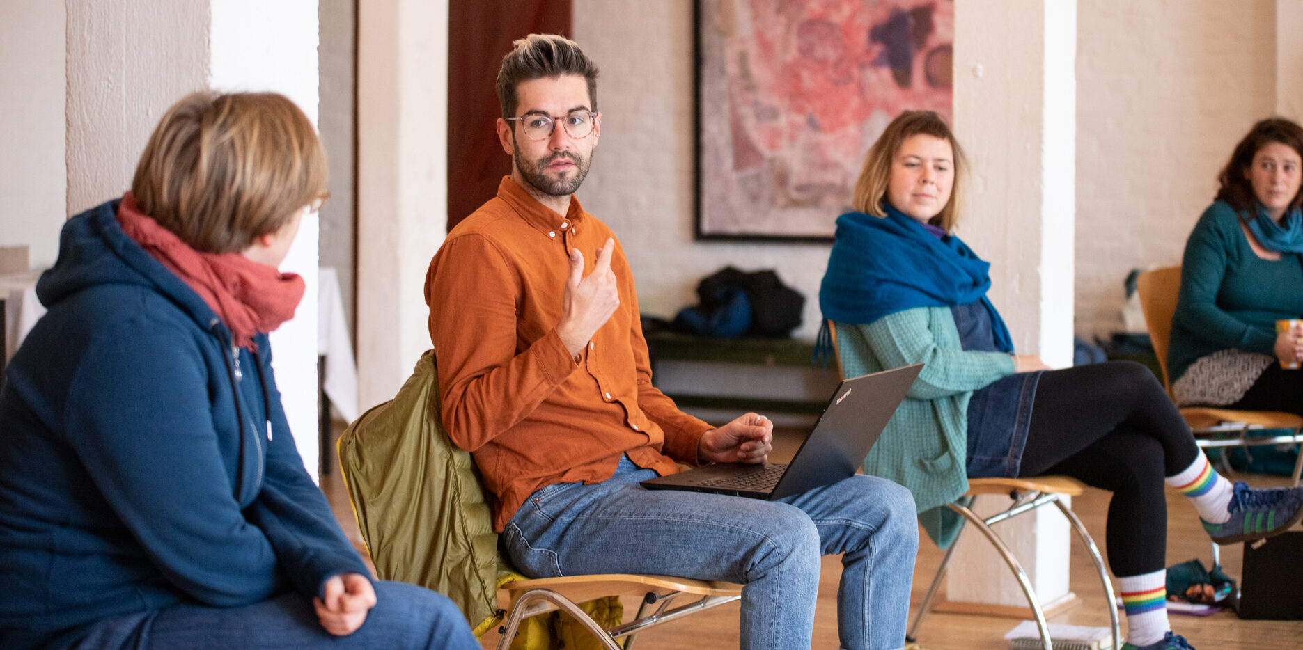 Jana, Simon, Evelyn und Marie vom F3_kollektiv sitzen nebeneinander in einem Stuhlkreis. Im Fokus des Bildes ist Simon mit einem Laptop auf dem Schoß. Er gestikuliert mit den Händen.