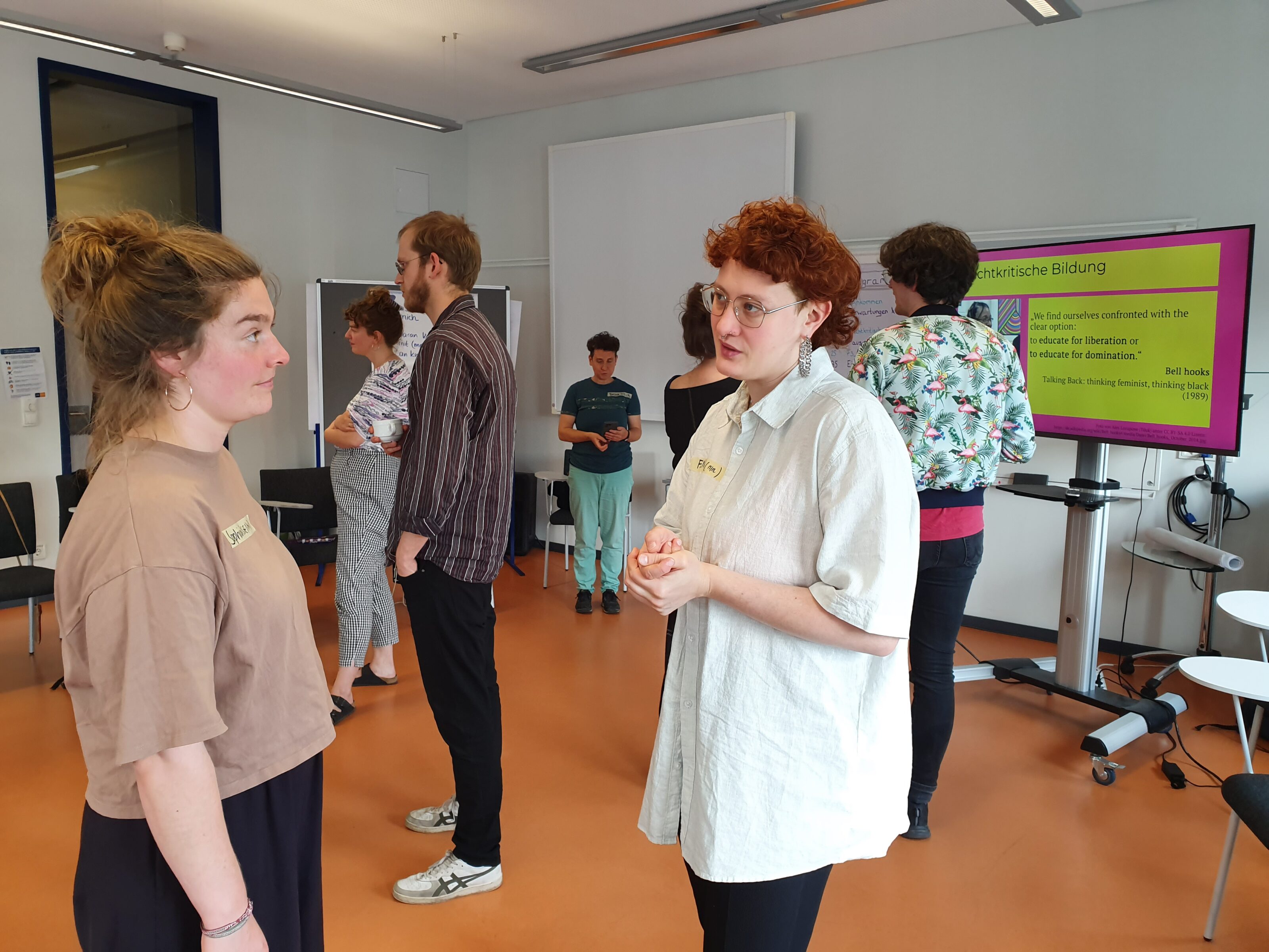 Teilnehmer*innen der Fortbildung stehen zu zweit im Raum verteilt und unterhalten sich. Im Hintergrund Präsentation zu Machtkritischer Bildung