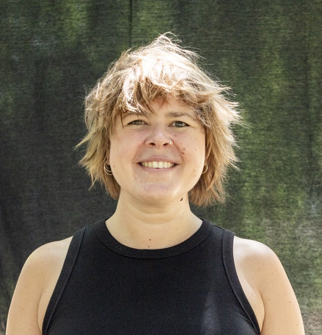 Portrait von Kollektivmitglied Katrin vor schwarzem Hintergrund. Sie trägt ein schwarzes Oberteil und lächelt in die Kamera.